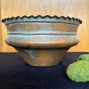 May include: A round copper bowl with a wide, flared body and a scalloped, wavy rim. The metal surface displays a weathered, brownish-orange patina with visible areas of green oxidation throughout the exterior, featuring a horizontal ridge.