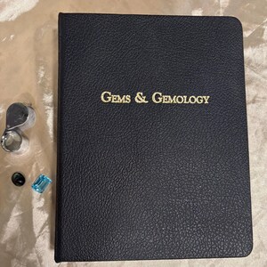 Puede incluir: Se muestra un libro negro texturizado con el texto dorado "Gems & Gemology". Una lupa, una gema negra y una gema azul están al lado del libro. El libro y las gemas están sobre una superficie de color claro.