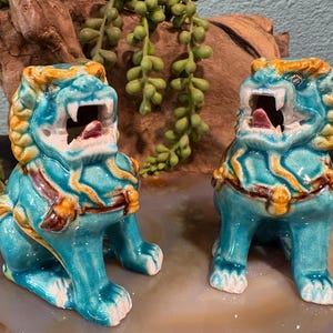 May include: Two turquoise ceramic Foo Dog statues with open mouths, showing their pink tongues. The statues have yellow and brown accents, and are sitting on a decorative surface. The background includes greenery and a piece of wood.