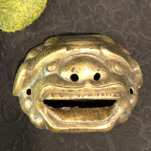 May include: A brass lion mask with a detailed design. The mask has a wide open mouth with sharp teeth and two holes for eyes.