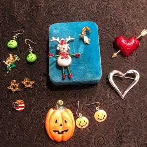 May include: Assortment of enamel pins and earrings featuring various holiday and seasonal themes, including a reindeer, a pumpkin, a heart, a star, a smiling face, and an angel. The pins and earrings are displayed on a black fabric background.