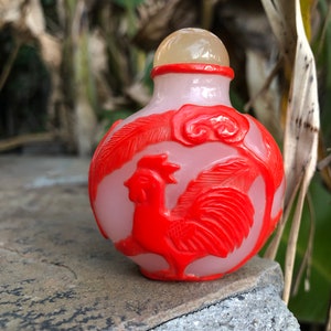 May include: A small, white, hand-carved glass bottle with a red rooster design. The bottle has a clear glass stopper.
