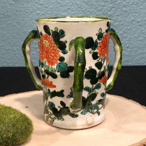 Puede incluir: Una taza de cerámica blanca con asas de estilo bambú verde y diseños florales en tonos verdes, naranjas y blancos.