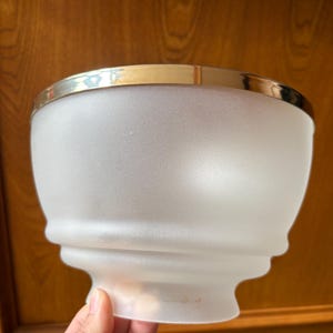 May include: A frosted glass bowl-shaped light fixture shade with a smooth, rounded silhouette and a wide, polished metallic gold-tone rim at the top. The glass has a matte, translucent texture that diffuses light, featuring a stepped base design.