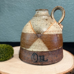 Op de afbeelding: Een kleine, bruine en witte keramische oliefles met een handvat. De fles heeft een bruine glazuur met witte strepen en het woord "Olie" is op de bodem gegraveerd.