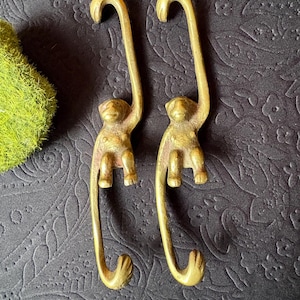 May include: Two identical brass monkey-shaped hooks, each featuring a monkey figure suspended by one arm from a curved top hook, with a second curved hook extending from the monkey's feet, creating a continuous vertical metal structure.