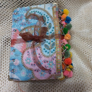 May include: A handmade journal with a colorful floral fabric cover and a decorative tassel trim. The journal has a blue and pink floral pattern with gold accents. The tassel trim is made of colorful beads and pom-poms.