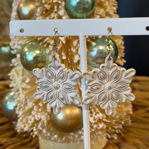 May include: A pair of white snowflake earrings with detailed patterns. The earrings are shown on a white stand, set against a small, decorated Christmas tree with gold and green baubles.