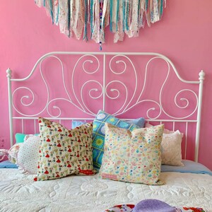 May include: A white metal bed frame with a pink wall behind it. The bed is made with a white quilt and has several colorful pillows on top. A purple hat and a floral patterned scarf are on the bed.