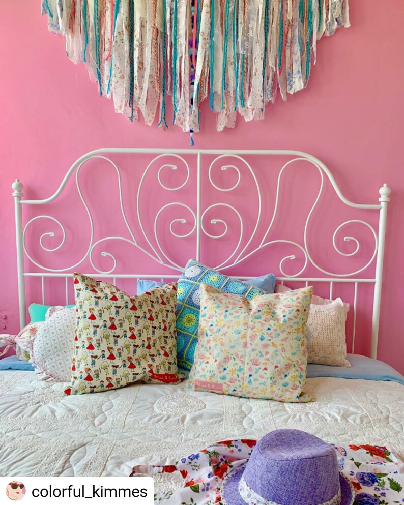 May include: A white metal bed frame with a pink wall behind it. The bed is made with a white quilt and has several colorful pillows on top. A purple hat and a floral patterned scarf are on the bed.