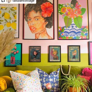 May include: A green velvet sofa with yellow, blue, and white patterned pillows. The sofa is in front of a wall with a pink and green color scheme. There are several framed prints on the wall, including a portrait of a woman with a red flower in her hair, a floral print, and three prints with the text "Lose Your Mind, Find Your Soul" and "Be Kind, To Your Mind".