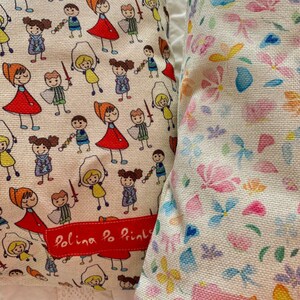 May include: Two white pillow covers with different patterns. One cover features a repeating pattern of colorful stick figures playing with toys. The other cover features a repeating pattern of colorful flowers and butterflies. Both covers have a red label that says "Polina Po Prints".
