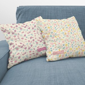 May include: Two decorative throw pillows with a floral pattern in shades of pink, yellow, and blue on a light blue couch. The pillows have a white background and the text "Bloom & Blush" is printed on the bottom of each pillow.