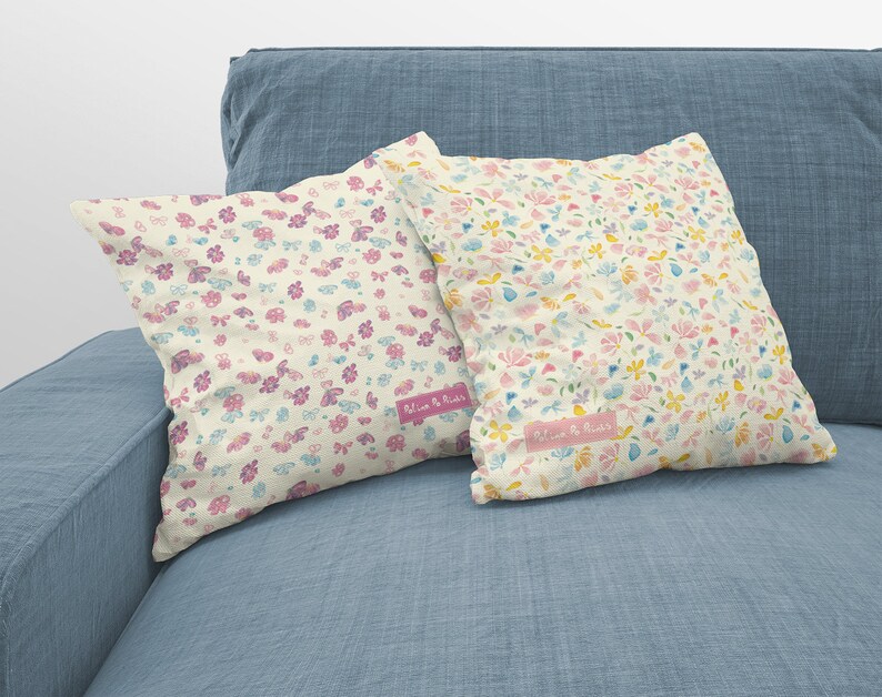 May include: Two decorative throw pillows with a floral pattern in shades of pink, yellow, and blue on a light blue couch. The pillows have a white background and the text "Bloom & Blush" is printed on the bottom of each pillow.