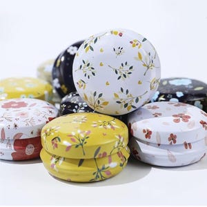 May include: A collection of small, round tins with floral patterns. The tins are in various colors, including white, yellow, and black, and feature designs of flowers and leaves. The lids are domed, and the tins appear to be made of metal.