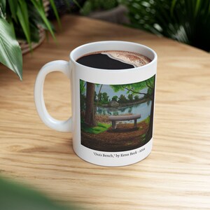 May include: White ceramic coffee mug with a landscape painting of a park bench overlooking a lake. The mug contains dark coffee. The painting is signed "'Ora's Bench,' by Kenn Beck - 2024."