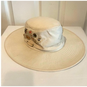 May include: A cream-colored canvas bucket hat with a wide, flat brim and a cylindrical crown. The hat features several small, decorative metal pins attached to the side of the crown, along with a metal eyelet and a fabric loop detail near the base.