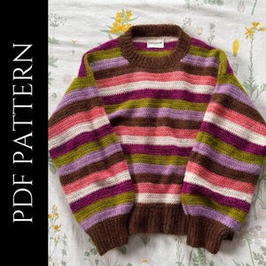 May include: A colorful, striped sweater with a brown collar, cuffs, and hem. The sweater features horizontal stripes in shades of brown, pink, purple, green, and white. The text "PDF PATTERN" is printed vertically on the left side of the image.