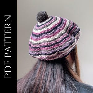May include: A handmade, striped crochet beret in shades of pink, purple, gray, and white, topped with a gray pom-pom. The text "PDF PATTERN" is vertically displayed on the left side of the image. The beret is shown from the back.