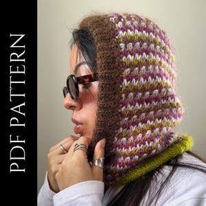 Może przedstawiać: Dzianinowy kaptur w odcieniach brązu, fioletu, zieleni i bieli. Kaptur ma wzorzystą fakturę i zielone wykończenie wokół twarzy. Tekst "PDF PATTERN" znajduje się po lewej stronie obrazu.