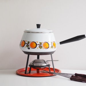 Op de afbeelding: Witte fonduepan met oranje en groen fruitontwerp, zwarte handgreep en een rode metalen basis. De pan staat op een wit oppervlak met zes fonduevorken.