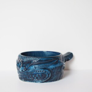 May include: A blue ceramic serving dish with a handle. The dish has a detailed design of a face with a wide mouth and sharp teeth.