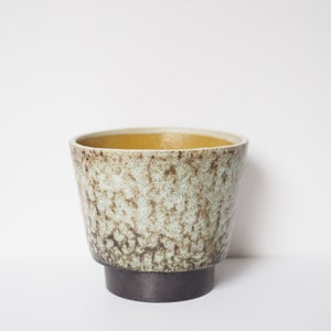 Op de afbeelding: Een keramische plantenpot met een gespikkeld groen glazuur en een gele rand. De pot heeft een brede basis en een smalle opening.