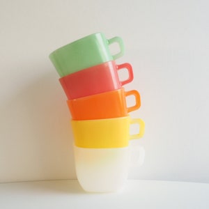 May include: A stack of five brightly coloured mugs in shades of green, red, orange, yellow, and white. Each mug has a square handle and a rounded body. The mugs are stacked at a slight angle against a white background.