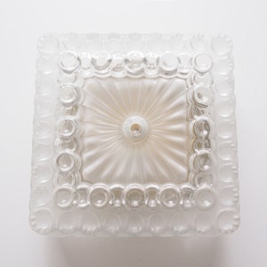 Op de afbeelding: Een vierkante plafondlamp van helder glas met een decoratief, getextureerd ontwerp. De lamp heeft een centrale koepel met een ronde opening in het midden.