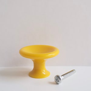 Op de afbeelding: Een gele plastic kastknop met een ronde bovenkant en een korte, ronde basis. Een zilveren schroef is inbegrepen.