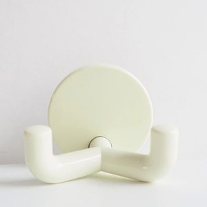 以下が含まれることがあります： 壁に物を掛けるための、2本の腕が付いた白いプラスチック製のフック。フックは丸いベースを持ち、接着剤で壁に取り付けられています。