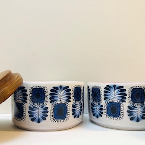 Op de afbeelding: Twee witte keramische voorraadpotten met een herhalend blauw bloemmotief van gestileerde vierkante en bladvormige figuren. Elke pot heeft een rond houten deksel, waarbij één deksel schuin tegen de zijkant van de pot aan rust.
