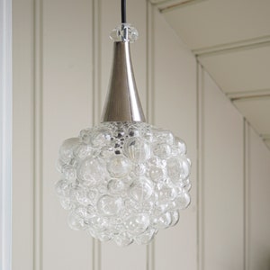 May include: A pendant light fitting featuring a spherical shade composed of numerous clear glass bubbles. The shade is attached to a smooth, conical, metallic silver-toned top piece that connects to a black cord extending upwards from the fitting.