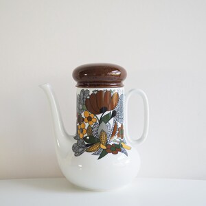 Peut inclure: Une cafetière en céramique blanche avec un couvercle brun et un motif floral. Les fleurs sont jaunes, orange, brunes et blanches. La cafetière a un bec verseur et une poignée.
