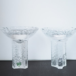 Op de afbeelding: Twee heldere glazen kaarsenstandaards met een getextureerd oppervlak. De standaards hebben een brede, ondiepe kom en een hoge, vierkante basis. De basis van elke standaard heeft een groen logo met de tekst "Orrefors Sweden".