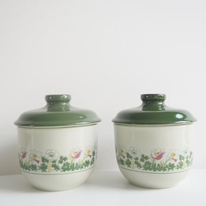 Op de afbeelding: Twee keramische potten met groene deksels en bloemenmotieven. De potten zijn wit met een groen en wit bloemenmotief rond het midden.