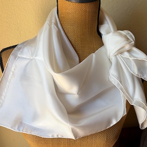 May include: A solid white square scarf with a smooth, lustrous finish draped around a mannequin. The scarf features a clean, hemmed edge and is tied in a simple knot on one side, creating soft folds and a draped appearance across the neck area.