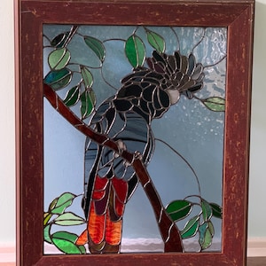May include: Stained glass artwork of a black cockatoo perched on a branch with green leaves. The bird has red and orange tail feathers and a gray crest. The artwork is framed in a dark brown wooden frame.