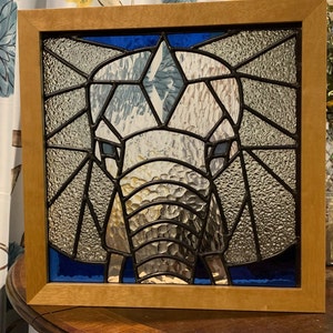 Può includere: Una finestra in vetro colorato che raffigura il viso di un elefante in tonalità di grigio e blu. La proboscide dell'elefante è visibile e lo sfondo è blu scuro.