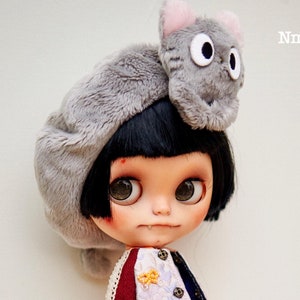 May include: A doll with black hair wearing a gray cat-shaped hat, a white and blue patterned jacket, a blue skirt, and blue and white sneakers. The doll has blue eyes and a light pink blush on her cheeks. The doll is standing on a wooden surface. "Nm Workshop" is written in the top right corner of the image.