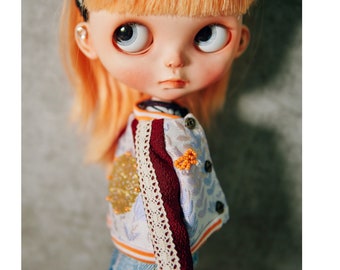 May include: A doll with blonde hair wearing a white and blue jacket with a red trim and a white lace detail. The jacket has a gold and orange embroidered design. The doll is wearing blue jeans and has a black headband with white flowers.