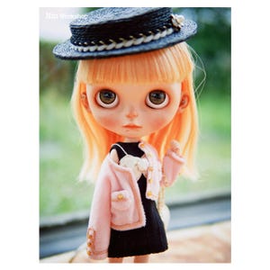 May include: A doll with large eyes, wearing a black dress, a pink jacket, and a black hat with a white flower. The doll has light orange hair and is set against a blurred green background. The text "Nm Workshop" is visible in the top left corner.
