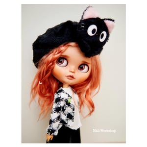 May include: A doll with long, wavy, pink hair wearing a black beret with a black cat plush on top. The doll has large, brown eyes and is wearing a black and white checkered jacket.