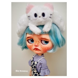May include: A doll with blue hair and a white cat hat. The doll has freckles and is wearing a purple shirt and a blue overall dress with white flowers.