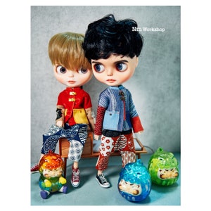 Può includere: Due bambole vestite con abiti di ispirazione asiatica tradizionale. La bambola a sinistra indossa una giacca rossa e gialla con pantaloni a fantasia blu e bianca. La bambola a destra indossa una giacca blu e bianca con pantaloni a fantasia rossa e bianca. Entrambe le bambole tengono in mano piccole figurine di animali colorate.