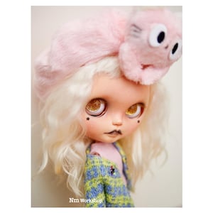 May include: A doll with blonde curly hair wearing a pink fuzzy hat with cat ears and a green and blue plaid jacket. The doll has large, sparkly eyes and a painted-on black mouth.