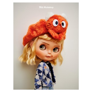 May include: A doll with blonde hair wearing a bright orange beret with a fuzzy orange animal face on top. The doll is wearing a blue and white checkered jacket.