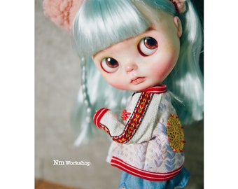 May include: A doll with blue hair and a white and red jacket with floral embroidery. The doll is wearing a blue skirt and has a pink headband with cat ears.