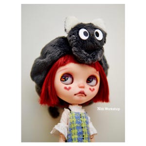 May include: A red-haired doll wearing a gray beret with a black cat plush on top. The doll has large blue eyes with black and orange details and pink hearts painted on her cheeks. The doll is wearing a white shirt with a plaid pinafore dress.