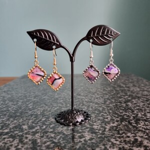 May include: Three pairs of dangle earrings with square-shaped resin insets. The resin is translucent and features swirls of pink, purple, and white. The earrings are displayed on a black metal stand.